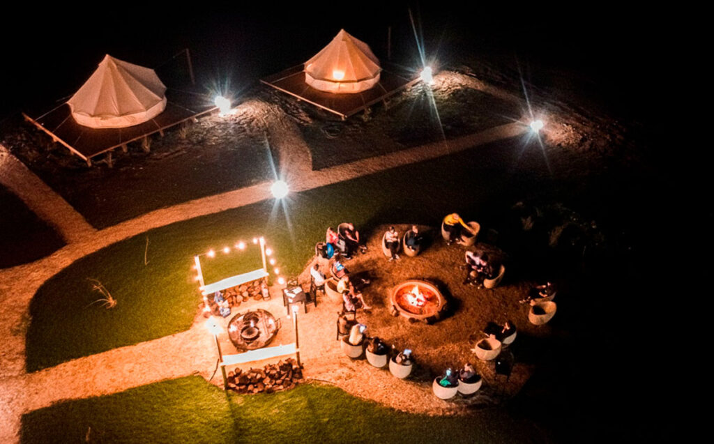 Vista aérea de noche del área de fogatas y carpas iluminadas en Terranova Glamping, con gente reunida.