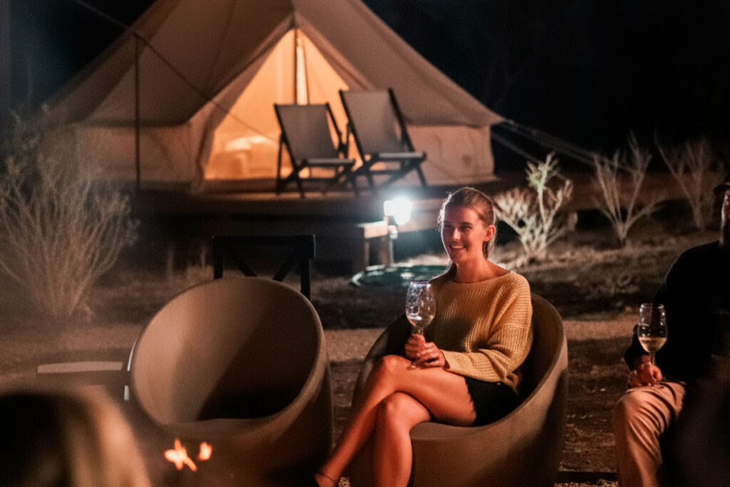 Mujer sonriendo y disfrutando de una copa de vino junto a la fogata en el área social de Terranova Glamping por la noche.