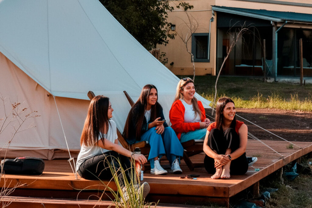 Cuatro compañeras de trabajo relajándose y charlando en la terraza de una carpa de glamping durante un evento de team building.